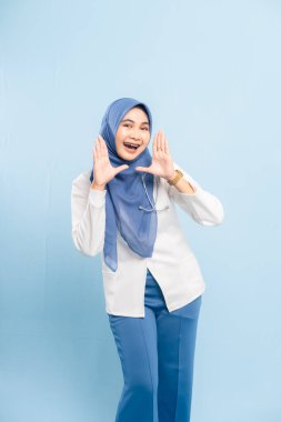 Heyecanlı bir Asyalı Müslüman doktor ya da hemşirenin portresi bağırıyor ya da yüksek sesle anons ediyor, el ele boğuşuyor, mavi arka planda izole edilmiş..