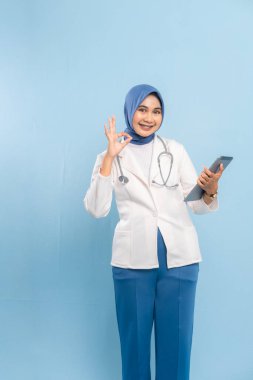 Tablet ve Başparmağıyla Asyalı Müslüman Doktor, Onaylama ve Dijital Sağlık Sinyali. Mavi arkaplanda izole edilmiş