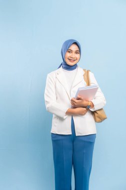 Gülümseyen ve kendinden emin genç bayan tıp öğrencisi ya da tesettürdeki doktor. Mavi arkaplanda izole edilmiş.