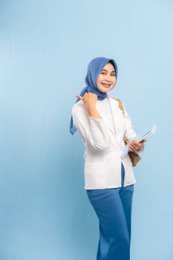 Gülümseyen ve kendinden emin genç Müslüman kız öğrenci mavi arka planda izole edilmiş önemli bir şeye işaret ediyor..