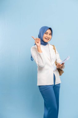 Gülümseyen ve kendinden emin genç Müslüman kız öğrenci mavi arka planda izole edilmiş önemli bir şeye işaret ediyor..