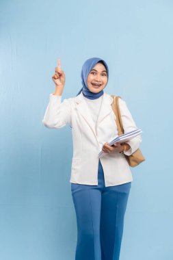 Gülümseyen ve kendinden emin genç Müslüman kız öğrenci mavi arka planda izole edilmiş önemli bir şeye işaret ediyor..
