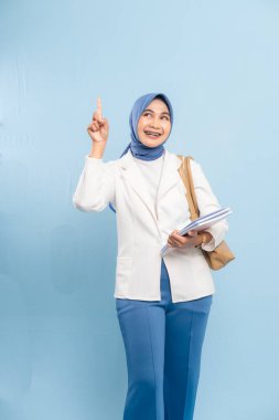 Gülümseyen ve kendinden emin genç Müslüman kız öğrenci mavi arka planda izole edilmiş önemli bir şeye işaret ediyor..