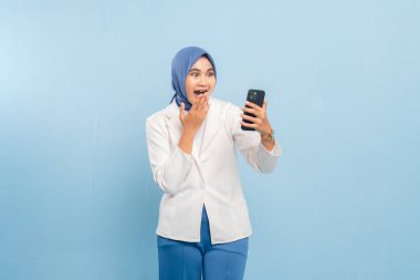 Genç Asyalı Müslüman iş kadını ya da telefonuna bakan öğrenci şaşırdı ya da şok oldu. Mavi bir arkaplanda izole edilmiş