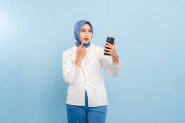 Genç Asyalı Müslüman iş kadını ya da telefonuna bakan öğrenci şaşırdı ya da şok oldu. Mavi bir arkaplanda izole edilmiş