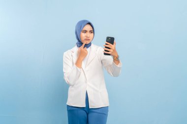 Düşünceli ya da kafası karışmış Asyalı genç Müslüman ya da elinde telefon tutarken bir fikri düşünen öğrenci. Mavi bir arkaplanda izole edilmiş.