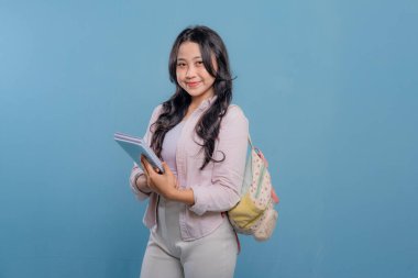 Bir kadın bir kitap ve bir sırt çantası tutuyor. Gülümsüyor ve kameraya bakıyor. Kendine güven ve hazır olma kavramı, çünkü kadın önümüzdeki güne hazır.