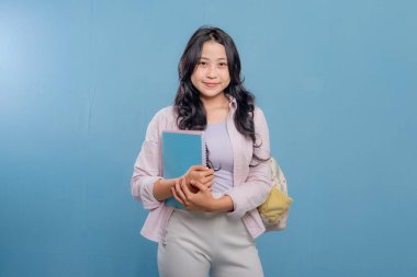 Bir kadın bir kitap ve bir sırt çantası tutuyor. Gülümsüyor ve kameraya bakıyor. Kendine güven ve hazır olma kavramı, çünkü kadın önümüzdeki güne hazır.