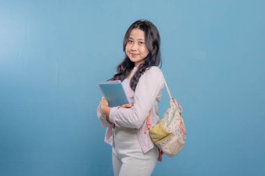 Bir kadın bir kitap ve bir sırt çantası tutuyor. Gülümsüyor ve kameraya bakıyor. Kendine güven ve hazır olma kavramı, çünkü kadın önümüzdeki güne hazır.