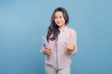 Bir kadın elinde mavi bir kitap tutuyor ve önemli bir şeye işaret ediyor. Gülümsüyor ve mutlu.