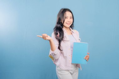 Bir kadın elinde mavi bir kitap tutuyor ve önemli bir şeye işaret ediyor. Gülümsüyor ve mutlu.