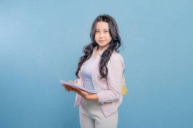 Bir kadın bir kitap ve bir sırt çantası tutuyor. Gülümsüyor ve kameraya bakıyor. Kendine güven ve hazır olma kavramı, çünkü kadın önümüzdeki güne hazır.