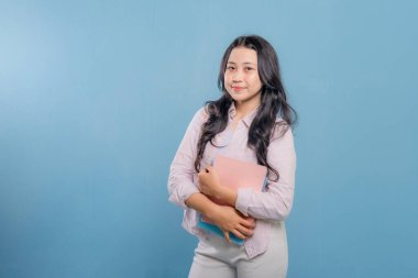 Bir kadın bir kitap ve bir sırt çantası tutuyor. Gülümsüyor ve kameraya bakıyor. Kendine güven ve hazır olma kavramı, çünkü kadın önümüzdeki güne hazır.