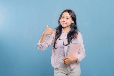 Bir kadın gülümsüyor ve elinde bir yığın kitap tutarken başparmağını kaldırıyor. Kadın akademik başarılarıyla gurur duyduğu için, başarı ve pozitiflik kavramı.