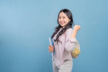 Bir kadın bir kitap ve bir sırt çantası tutuyor. Gülümsüyor ve kameraya bakıyor. Kendine güven ve hazır olma kavramı, çünkü kadın önümüzdeki güne hazır.