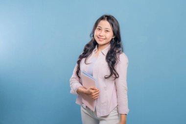 Bir kadın bir kitap ve bir sırt çantası tutuyor. Gülümsüyor ve kameraya bakıyor. Kendine güven ve hazır olma kavramı, çünkü kadın önümüzdeki güne hazır.