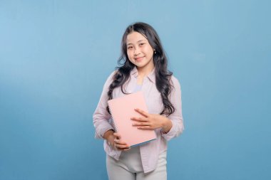 Bir kadın bir kitap ve bir sırt çantası tutuyor. Gülümsüyor ve kameraya bakıyor. Kendine güven ve hazır olma kavramı, çünkü kadın önümüzdeki güne hazır.