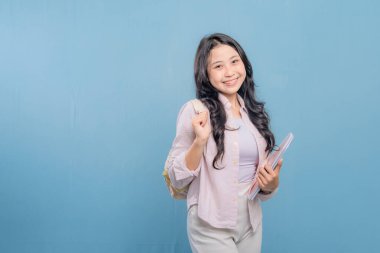 Bir kadın bir kitap ve bir sırt çantası tutuyor. Gülümsüyor ve kameraya bakıyor. Kendine güven ve hazır olma kavramı, çünkü kadın önümüzdeki güne hazır.