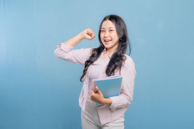 Bir kadın bir kitap ve bir sırt çantası tutuyor. Gülümsüyor ve kameraya bakıyor. Kendine güven ve hazır olma kavramı, çünkü kadın önümüzdeki güne hazır.