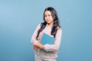 Bir kadın elinde mavi bir defter tutuyor ve kameraya bakıyor. Kafası karışık ya da bir şeyden emin değil.