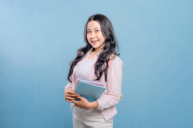 Bir kadın bir kitap ve bir sırt çantası tutuyor. Gülümsüyor ve kameraya bakıyor. Kendine güven ve hazır olma kavramı, çünkü kadın önümüzdeki güne hazır.