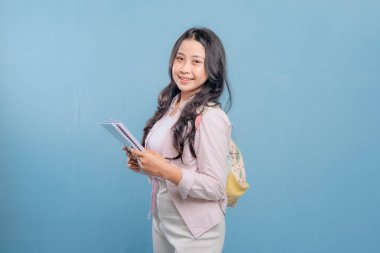 Bir kadın bir kitap ve bir sırt çantası tutuyor. Gülümsüyor ve kameraya bakıyor. Kendine güven ve hazır olma kavramı, çünkü kadın önümüzdeki güne hazır.