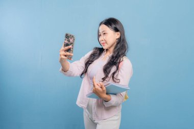 Bir kadın cep telefonuyla selfie çekiyor. O, pembe bir gömlek ve mavi pantolon giyiyor. Elinde mavi bir kitap ve pembe bir kitap tutuyor.