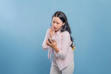 Mavi bir duvarın önünde duran bir kadın telefonuna bakıyor. Telefonundaki bir şey onu şaşırttı ya da şok etti.