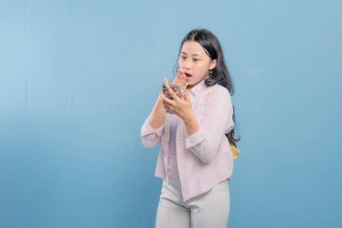 Mavi bir duvarın önünde duran bir kadın telefonuna bakıyor. Telefonundaki bir şey onu şaşırttı ya da şok etti.