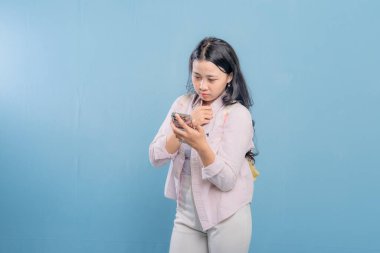 Mavi bir duvarın önünde duran bir kadın telefonuna bakıyor. Telefonundaki bir şey onu şaşırttı ya da şok etti.