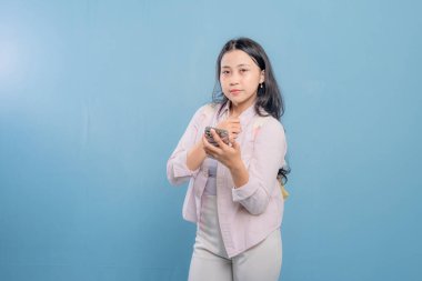 Mavi bir duvarın önünde duran bir kadın telefonuna bakıyor. Telefonundaki bir şey onu şaşırttı ya da şok etti.