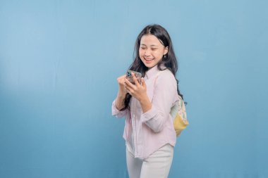 Mavi bir duvarın önünde duran bir kadın telefonuna bakıyor. Telefonundaki bir şeyle gülümsüyor ve mutlu.