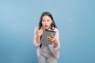 Bir kadın elinde bir tablet tutuyor ve ona şaşırmış bir ifadeyle bakıyor. Pembe gömlek ve beyaz pantolon giyiyor.