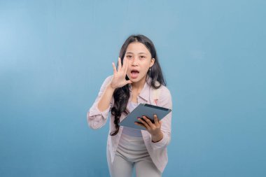 Bir kadın elinde bir tablet tutuyor ve yüksek sesle gürültü yapıyor. Pembe gömlek ve beyaz pantolon giyiyor.