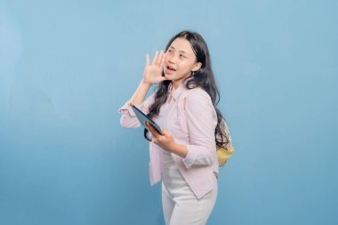 Bir kadın elinde bir tablet tutuyor ve yüksek sesle gürültü yapıyor. Pembe gömlek ve beyaz pantolon giyiyor.