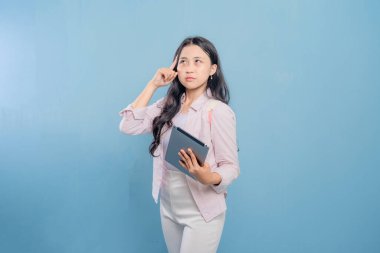 Mavi bir duvarın önünde başı yana eğilmiş bir kadın duruyor. Elinde bir tablet tutuyor.