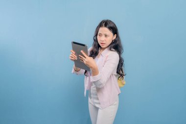 Bir kadın elinde bir tablet tutuyor ve ona şaşırmış bir ifadeyle bakıyor. Pembe gömlek ve beyaz pantolon giyiyor.