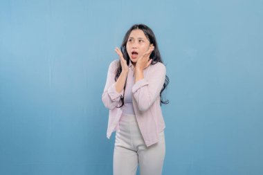 Uzun saçlı bir kadın mavi bir duvarın önünde duruyor. O, pembe bir gömlek ve beyaz pantolon giyiyor. O, ellerini yüzüne koydu ve başka yere bakıyor. Sürpriz ya da şok kavramı