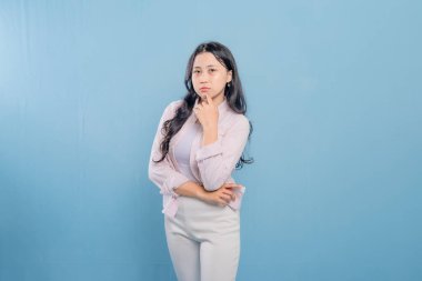 Mavi bir duvarın önünde pembe ceketli ve beyaz pantolonlu bir kadın duruyor. Aşağıya bakıyor ve derin düşüncelere dalmış.