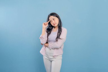 Mavi bir duvarın önünde pembe ceketli ve beyaz pantolonlu bir kadın duruyor. Aşağıya bakıyor ve derin düşüncelere dalmış.