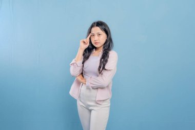 Mavi bir duvarın önünde kollarını kavuşturmuş bir kadın duruyor. Aşağıya bakıyor ve derin düşüncelere dalmış.