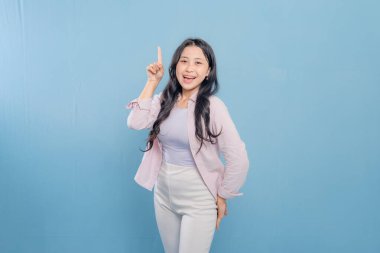 Mavi bir duvarın önünde pembe gömlekli ve beyaz pantolonlu bir kadın duruyor. Parmağını kaldırıp gülümsüyor.