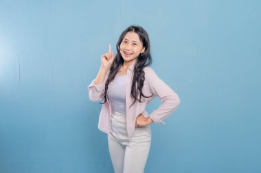 Mavi bir duvarın önünde pembe gömlekli ve beyaz pantolonlu bir kadın duruyor. Parmağını kaldırıp gülümsüyor.