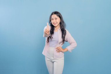 Mavi bir duvarın önünde pembe gömlekli ve beyaz pantolonlu bir kadın duruyor. Parmağını kaldırıp gülümsüyor.