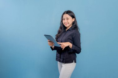 Bir kadın tablet tutuyor ve gülümsüyor. O, siyah bir gömlek ve beyaz pantolon giyiyor. Verimlilik ve odaklanma kavramı, kadının çalışma veya öğrenim için tableti kullanması gibi