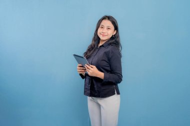 Bir kadın gülümsüyor ve elinde bir tablet tutuyor. O, siyah bir gömlek ve beyaz pantolon giyiyor. Profesyonellik ve güven kavramı