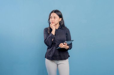 Bir kadın mavi bir duvarın önünde duruyor, elindeki tablete bakıyor. Derin düşüncelere dalmış, muhtemelen bir karar ya da sorun düşünüyor.