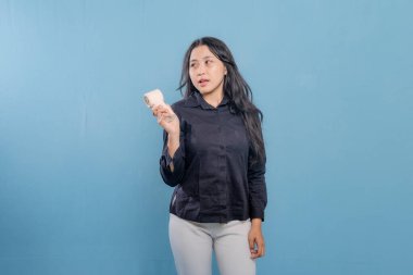 Bir kadın elinde bir cep telefonu tutuyor ve ona bakıyor. Siyah gömlek ve beyaz pantolon giyiyor.