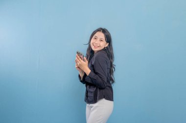 Mavi bir duvarın önünde duran bir kadın telefonuna bakıyor. Telefonundaki bir şeyle gülümsüyor ve mutlu.