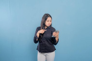 Mavi bir duvarın önünde duran bir kadın telefonuna bakıyor. Telefonundaki bir şey onu şaşırttı ya da şok etti.